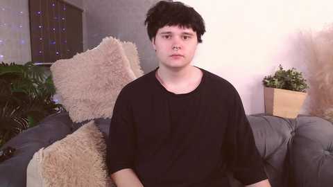 Media: A video of a young, pale-skinned man with short black hair wearing a black t-shirt, sitting on a gray couch. The background features a fluffy beige pillow, a wooden plant stand, and green foliage.