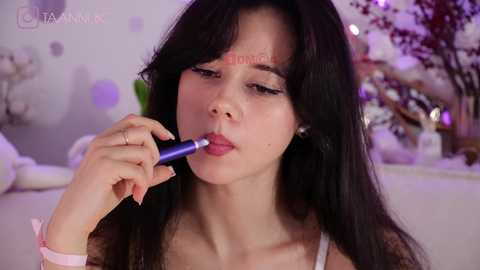 Media: Video of an Asian woman with long black hair, fair skin, and wearing a light pink top, applying lipstick. Background features a blurred, softly lit room with floral decor and a white teddy bear.