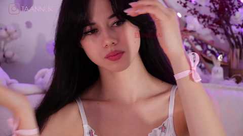 Media: Video of a young woman with long black hair, light skin, and a slender physique, wearing a white camisole with pink bows, sitting in a softly lit room with pastel decor.