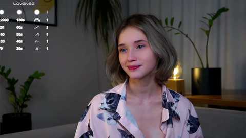 Media: Video of a young woman with shoulder-length blonde hair, wearing a pink floral blouse, standing indoors near a potted plant, dimly lit room with a TV showing \"LOVEHACK\" on the left.