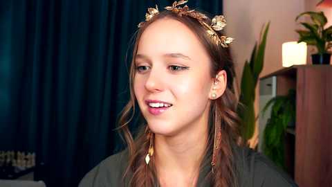 Media: Video of a young, smiling woman with light skin and brown hair in braids adorned with gold leaf crowns, wearing a green top, against a dark curtain background.