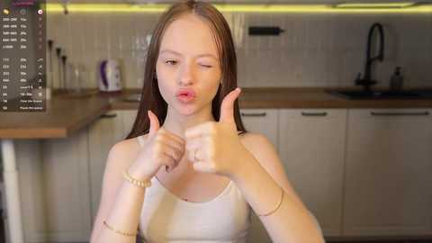 Media: Video of a young woman with fair skin, long brown hair, and a slim build, giving a thumbs-up in a modern kitchen with white cabinets and a wooden countertop.