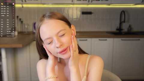 Media: Video of a fair-skinned woman with long brown hair, wearing a beige top, leaning back in a modern kitchen with white cabinets and wooden countertops, eyes closed, hands on her cheeks.