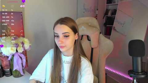 Media: Video of a young woman with long brown hair in a white t-shirt, seated in a room with a mannequin, a microphone stand, and a floral arrangement on a table.