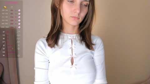 Media: Video of a young Caucasian woman with straight, medium-length brown hair, wearing a white, long-sleeve top with a small bow at the neckline. She looks down, expression neutral. Background shows a calendar with dates and a beige wall.