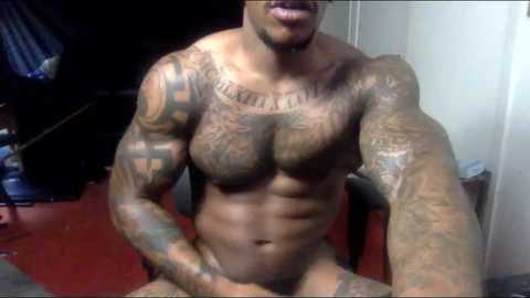 Media: Video of a muscular, tattooed black man with a goatee, wearing glasses, sitting shirtless with visible abs, arms, and chest tattoos. Background shows a red carpet and dimly lit room.