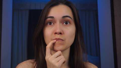 Media: Video of a young woman with long, straight brown hair, fair skin, and light brown eyes, sitting indoors with her fingers touching her lips, against a blurred, dark-blue and white background.