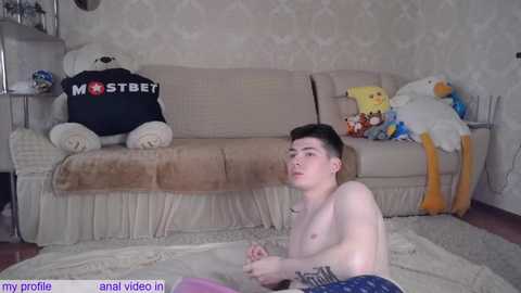 Media: A young, shirtless, fair-skinned man with short dark hair, wearing pajama pants, sits on a beige sofa, surrounded by plush toys in a cozy, patterned room.