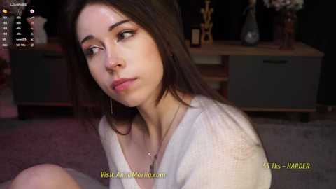 Media: A video of a young woman with long dark hair, fair skin, and a neutral expression, wearing a white robe. She is in a dimly lit room with a wooden cabinet in the background.