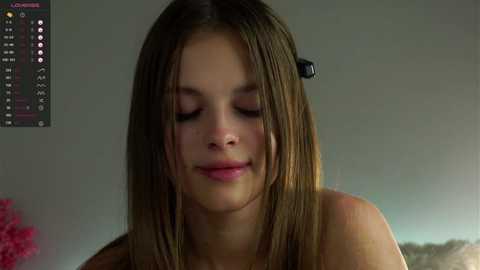 Media: Video of a young Caucasian woman with straight, shoulder-length brown hair, wearing a black tank top. She has fair skin and closed eyes, appearing calm and relaxed. Background shows a gray wall with a pink plant and a digital clock.