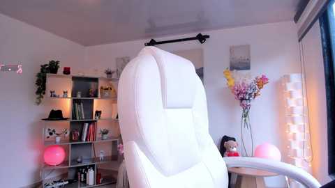 Media: Video of a modern, white leather recliner chair in a brightly lit, minimalist living room with white walls, bookshelves, potted plants, and decorative flowers.