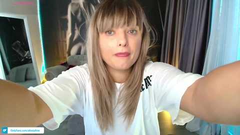 Media: Video of a young woman with straight, light brown hair, fair skin, and a neutral expression. She wears a white t-shirt and stands in a dimly lit room with a large black-and-white poster on the wall.