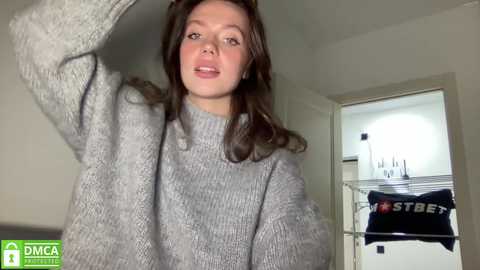 Media: Video of a young Caucasian woman with fair skin, brown hair, and a slim build, wearing a gray knit sweater. She stands in a modern, bright room with white walls, a doorway, and a laundry rack holding a dark blue garment.