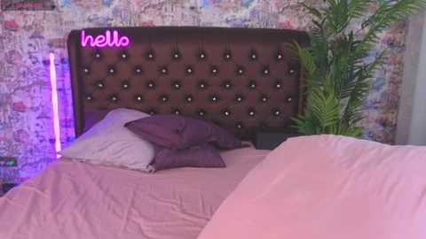 Media: Video of a modern bedroom with a tufted brown headboard, pink sheets, purple pillows, and a fern plant. Neon \"hello\" sign on the brick wall.