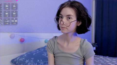 Video of a young woman with short brown hair, wearing glasses, a gray top, and sitting in a bedroom with blue walls, a bed with a blue patterned blanket, and a string of colorful lights.