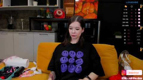 Media: Video of a young woman with straight brown hair, wearing a black T-shirt with a smiley face design, sitting on a yellow couch in a modern kitchen.