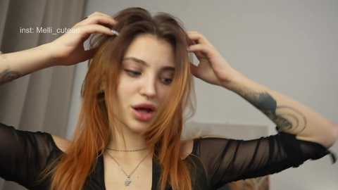 Media: Video of a young woman with fair skin, long brown hair, and multiple tattoos, adjusting her hair with her hands. She wears a sheer black top and a delicate necklace. Background is a neutral indoor setting.