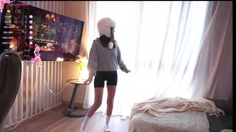 Media: Video of a young woman in a gray hoodie, black shorts, and white beanie, playing a video game near a large TV screen and a lit-up window in a cozy, sunlit room.