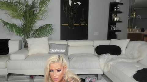 Media: A video of a blonde woman with long hair and dark makeup, sitting on a white sectional sofa in a modern, minimalist living room.