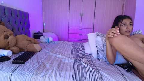 Media: A video of a young woman with medium brown skin and long black hair lying on a bed with striped sheets, holding her foot, in a room with purple lighting and wooden furniture.