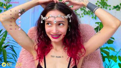 Media: Video of a young woman with long, wavy hair, fair skin, and tattoos, wearing a floral headband, black top, and red lipstick, seated in front of a pink, fluffy chair.