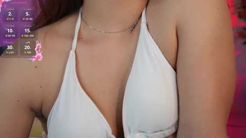 Media: Video of a woman wearing a white halter bikini top, revealing her cleavage. She has a thin silver necklace with a cross pendant. Background includes a digital timer display with numbers and pink floral design.