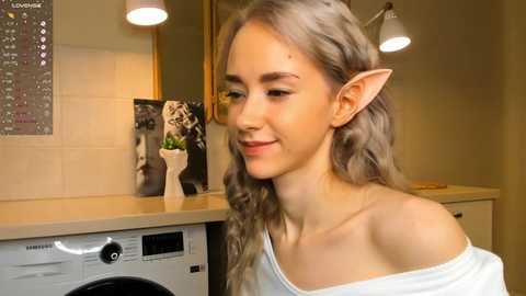 Media: Video of a young woman with long, wavy gray hair, pointy elf ears, and a white off-shoulder top, smiling in a beige-tiled kitchen with modern lighting and a framed black-and-white photo.