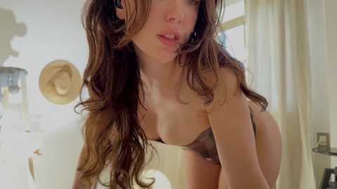 Media: A video of a brunette woman with long, wavy hair, wearing a black bra, leaning forward in a bright, softly lit room with white curtains and a wooden hat rack.