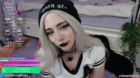 Media: A young woman with platinum blonde hair, dark lipstick, and a black beanie with \"Sith Lord\" text, wearing a black choker, sits on a purple carpeted floor in a bedroom.