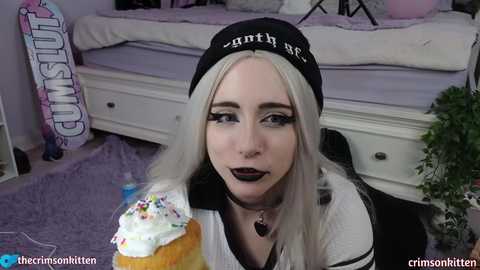 Media: Video of a young, fair-skinned woman with platinum blonde hair and dark makeup, wearing a black beanie, black choker, and black top. She holds a cupcake with colorful frosting. Background shows a bed, purple carpet, and a \"Cum Slut\" poster.