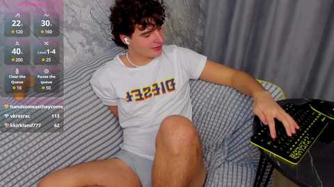 Media: Video of a young, fair-skinned man with curly brown hair, wearing a white T-shirt with \"FEB 21\" and grey shorts, sitting on a grey-striped couch, playing a video game on a neon-colored keyboard.