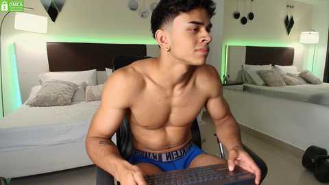 Media: Video of a shirtless, muscular, light-skinned Latino man with short, curly hair, wearing blue briefs, sitting at a desk in a modern, well-lit bedroom.