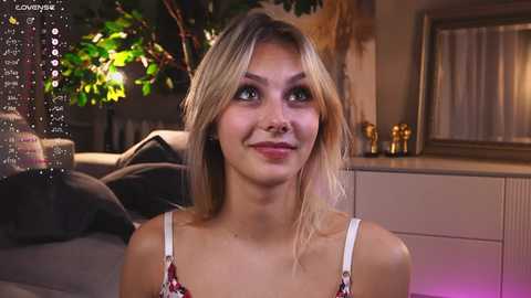 Media: Video of a smiling young woman with light skin, blonde hair, and green eyes, wearing a white and red floral-patterned spaghetti strap top. Background features a cozy living room with a fireplace, plants, and dim lighting.