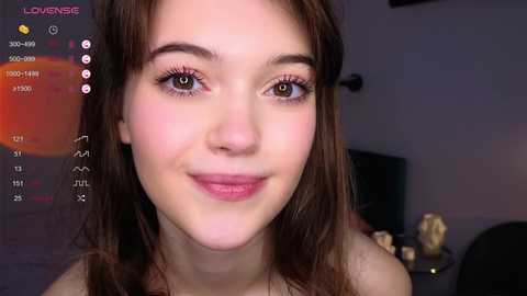 Media: Video of a young Caucasian woman with fair skin, brown hair, and green eyes, smiling softly. She wears a light-colored top. Background features a dimly lit room with a TV and blurred objects.