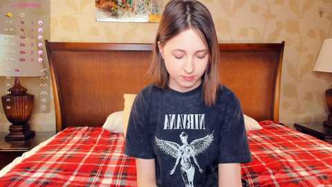 Media: Video of a young Caucasian woman with shoulder-length brown hair, wearing a black \"AC/DC\" t-shirt, sitting on a bed with a red plaid blanket in a cozy, wallpapered bedroom.