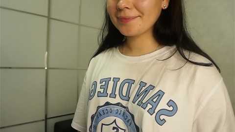 Media: Video of a smiling young woman with long dark hair, wearing a white San Diego State University T-shirt, standing in a tiled bathroom with white walls.