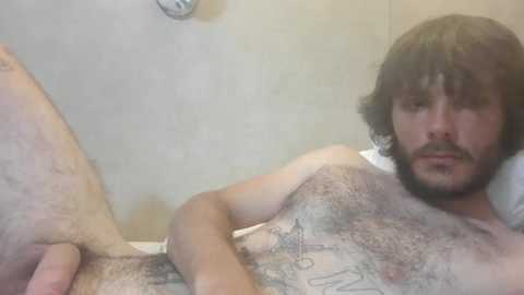 Media: Video of a bearded, light-skinned man with dark hair, lying naked on a bed with a tiled wall in the background, displaying a tattoo on his chest.