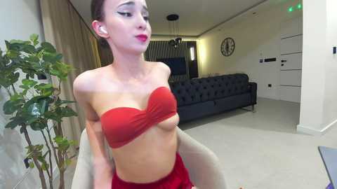 Media: Video of a slender, light-skinned woman with long brown hair, wearing a red strapless bandeau and red shorts, sitting in a beige chair in a modern living room with a black couch, green plants, and a circular clock.