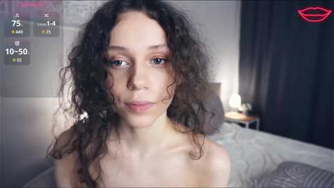 Media: Video of a young, light-skinned woman with curly black hair, blue eyes, and a slender physique, topless, in a dimly lit bedroom.