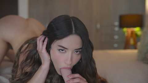 Media: Video of a nude, light-skinned woman with long dark hair performing oral sex on a man in a bedroom. Background shows wooden wardrobe and lamp.