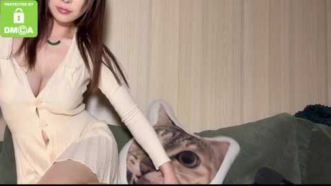 Media: Video of an East Asian woman with long brown hair, wearing a cream button-down dress, sitting on a green couch with a cat pillow, against a beige wall.