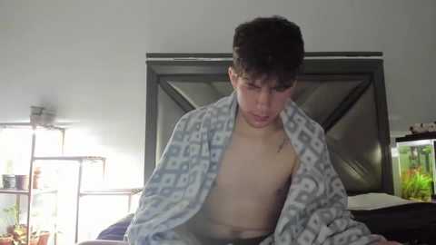 Media: Video of a young, shirtless boy with curly hair, wrapped in a patterned blanket, standing in a dimly lit room with a fish tank and shelves in the background.