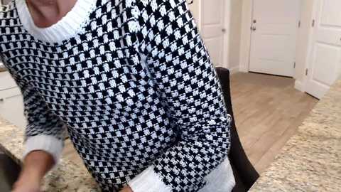 Media: Video of a person wearing a black-and-white herringbone-patterned sweater, standing indoors in a modern kitchen with light wood floors and white cabinetry.