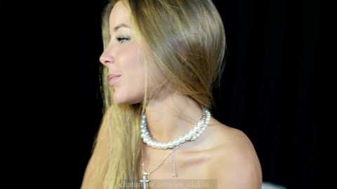 Media: A video of a young woman with long, straight blonde hair, wearing a pearl necklace, set against a black background. She has a fair complexion and is seen in profile.