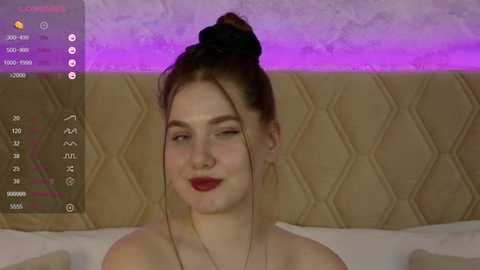 Media: Video of a fair-skinned woman with red lipstick, brown hair in a bun, on a beige tufted bed, wearing a gold necklace, smiling, against a purple-lit wall.