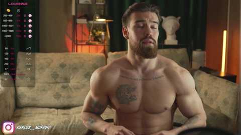 Media: Video of a muscular, shirtless man with a beard and tattoos, sitting on a floral sofa in a dimly lit living room.