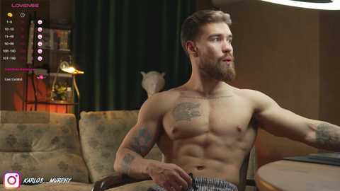 Media: Video of a shirtless, muscular man with tattoos, sitting in a cozy living room with a wooden table.