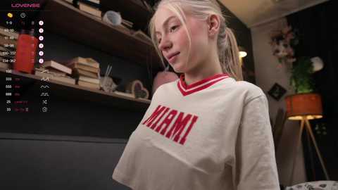 Media: Video of a young woman with fair skin, light blonde hair, wearing a \"MAMI\" sports jersey, in a modern, dimly lit room with bookshelves and potted plants.