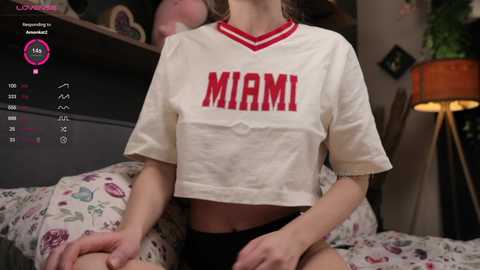 Media: Video of a young woman in a bedroom, wearing a cropped \"MAMI\" t-shirt, sitting on a bed with floral sheets. Background includes a lamp, plants, and a bookshelf.