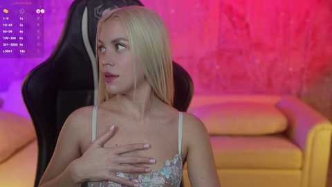 Media: Video of a blonde woman with fair skin, wearing a floral dress, sitting on a yellow couch, with a man's hand on her shoulder, in a room with purple and pink lights.